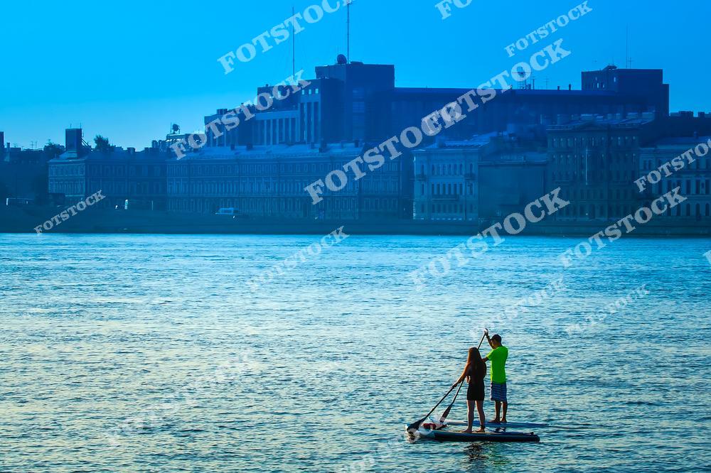 На фотографии двое человек  катаются на сапборде по реке на фоне городских зданий. Вечерний или утренний свет и спокойная вода создают атмосферу умиротворения и гармонии с природой даже в пределах города. Подходит для тематики активного отдыха, водных развлечений и городских пейзажей.